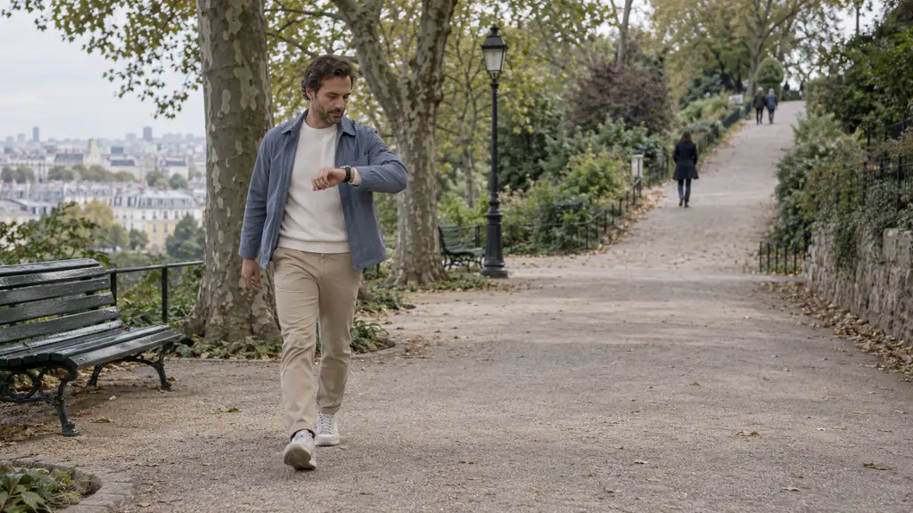 Editorial photograph of an adult brisk-walking on a flat park path while checking a wristwatch, with a gentle uphill trail visible farther ahead, no text or logo